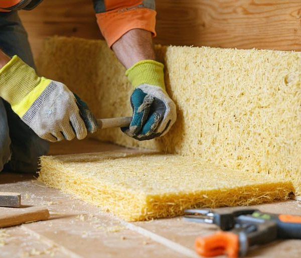 Gloved hands press thick hemp insulation batts into a wood-stud wall during a home renovation, with blurred tools, a mycelium panel sample, and a bale of sheep’s wool in the background.